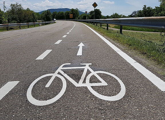 Eine Landstraße auf der ein Schutzstreifen für Radfahrende mit einer weißen gestrichelten Linie markiert ist.